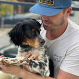 A man in a white shirt and the "Shop Hat | Trust Your Gut Vintage Slate" by Ballad of the Bird Dog holds a black and white dog outdoors, gently holding its front paws.