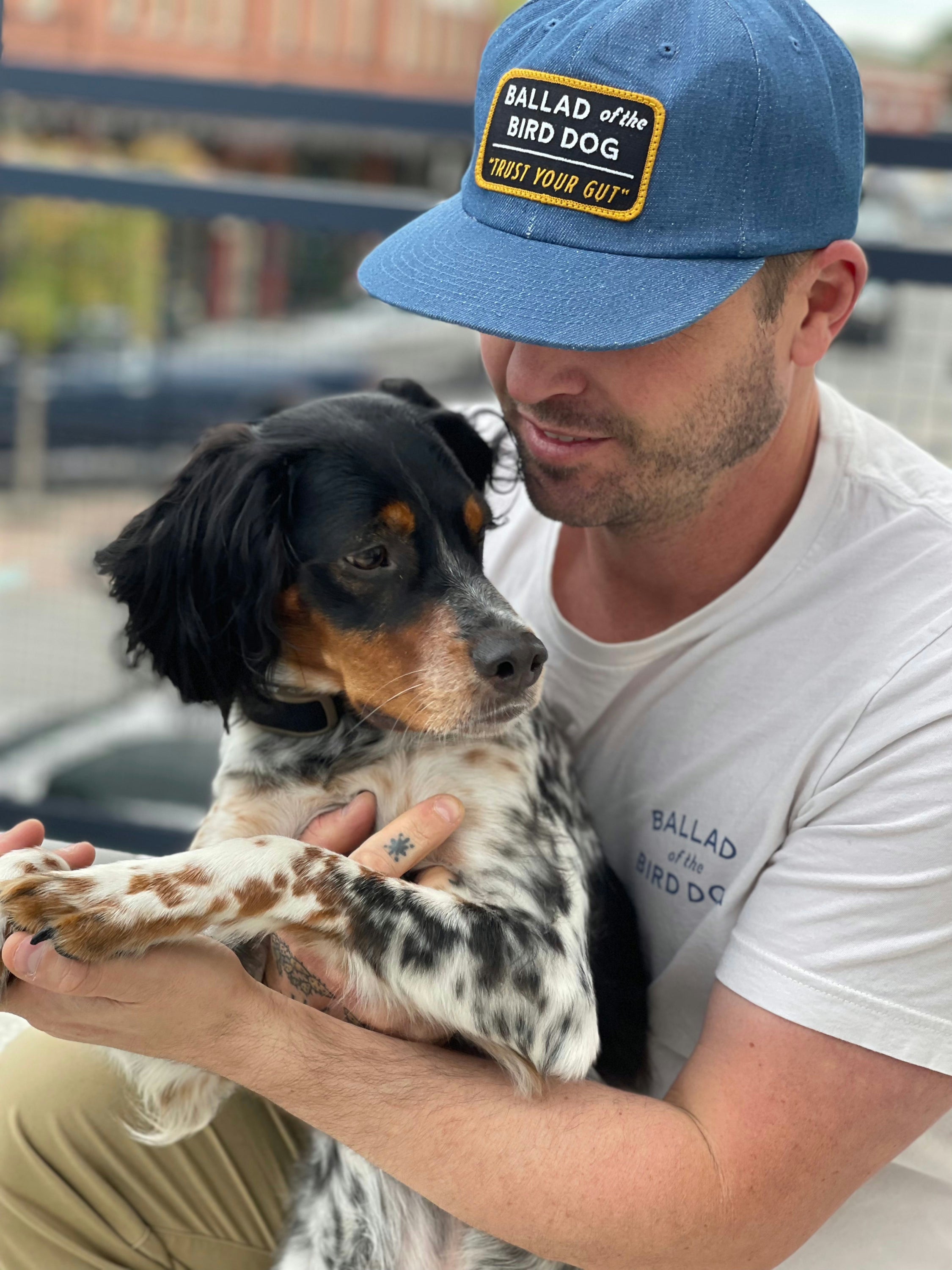 A man in a white shirt and the "Shop Hat | Trust Your Gut Vintage Slate" by Ballad of the Bird Dog holds a black and white dog outdoors, gently holding its front paws.