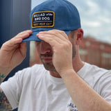 A man adjusts his Ballad of the Bird Dog "Shop Hat | Trust Your Gut Vintage Slate" and matching white T-shirt outdoors, with a blurred building in the background.