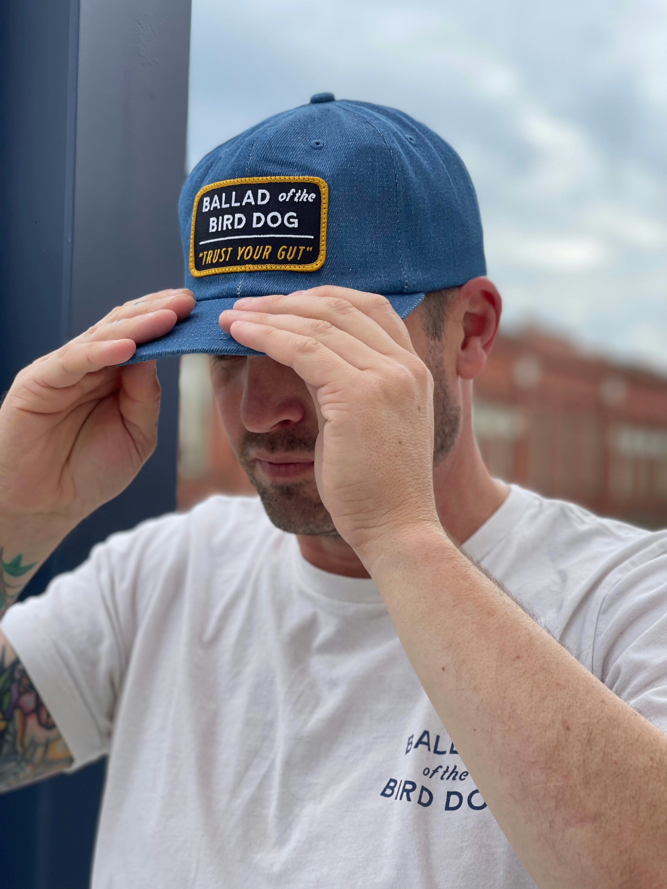 A man adjusts his Ballad of the Bird Dog "Shop Hat | Trust Your Gut Vintage Slate" and matching white T-shirt outdoors, with a blurred building in the background.