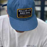 A person wears a Ballad of the Bird Dog “Shop Hat | Trust Your Gut Vintage Slate” with its patch, paired with a white shirt featuring the same brand name partly visible.