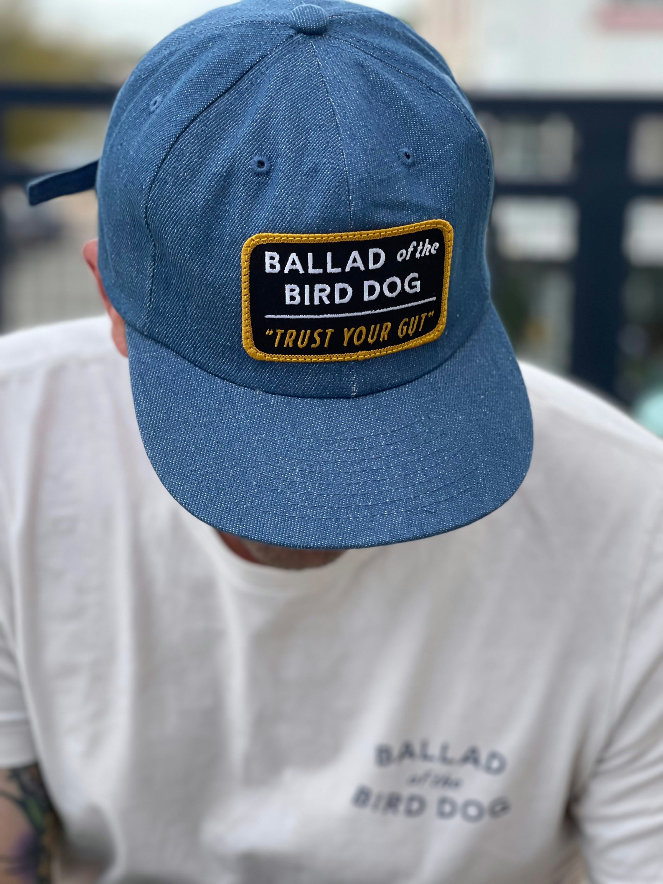 A person wears a Ballad of the Bird Dog “Shop Hat | Trust Your Gut Vintage Slate” with its patch, paired with a white shirt featuring the same brand name partly visible.