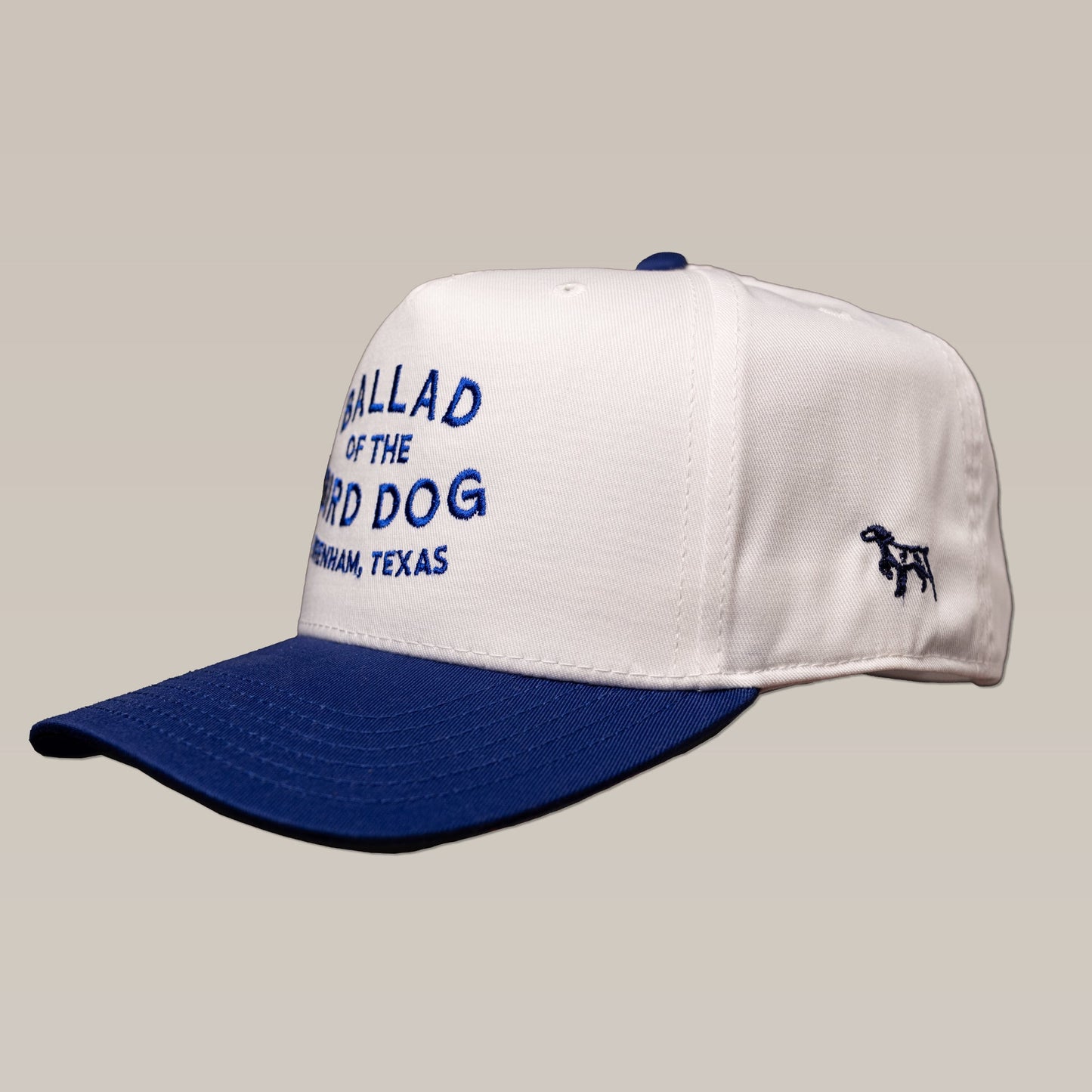 A white and blue Shop Hat by Ballad of the Bird Dog, featuring "Ballad of the Bird Dog Brenham, Texas" embroidery on the front and a small dog image on the side.