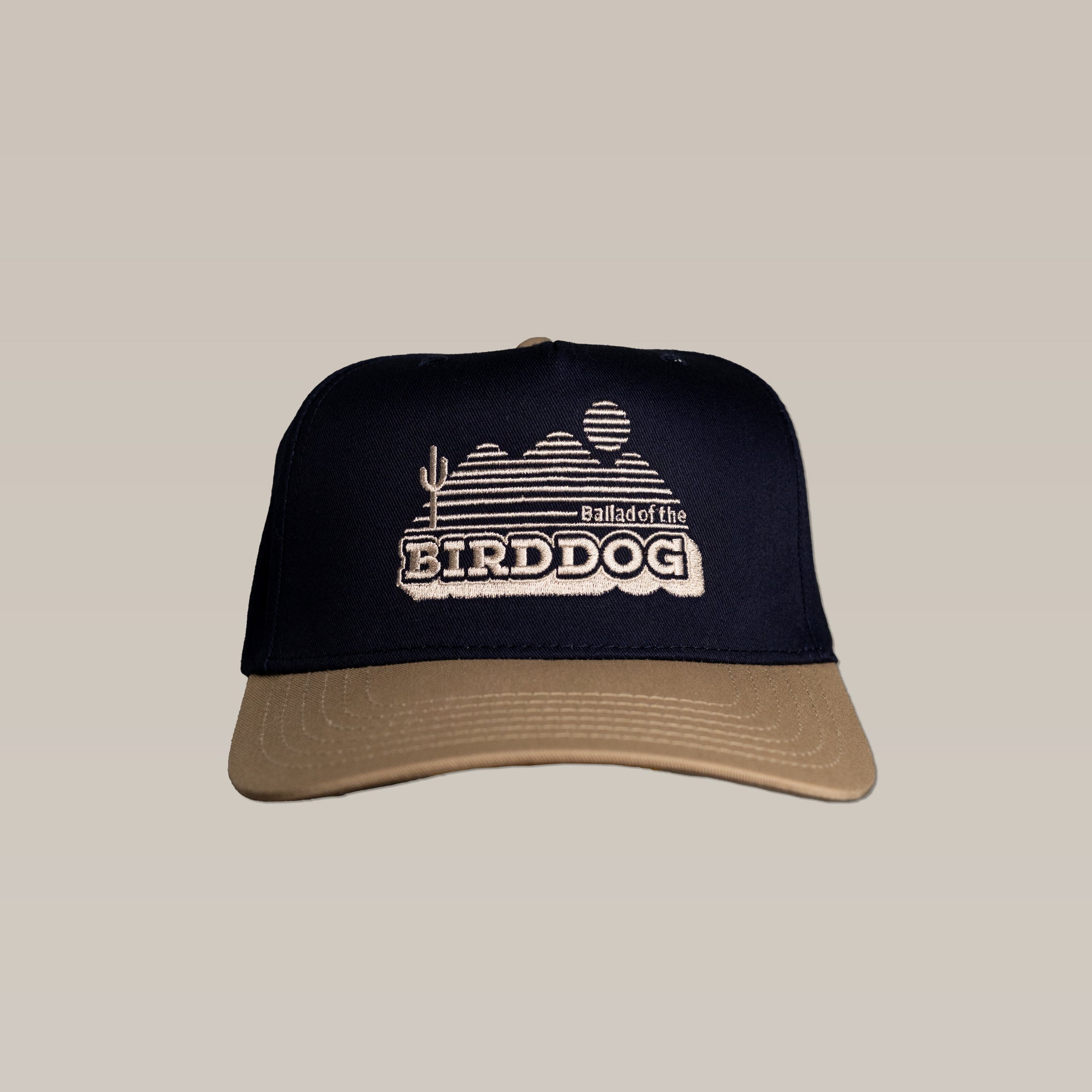 The Shop Hat by Ballad of the Bird Dog features a navy and tan design with "Ballad of the BIRDDOG" and a desert scene embroidered on the front, inspired by vintage bird dog hats.