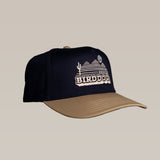 The Shop Hat by Ballad of the Bird Dog is a navy blue and tan cap featuring a desert landscape and "BIRDDOG" embroidered on the front, inspired by classic vintage logo style and the Vintage Bird Dog design.