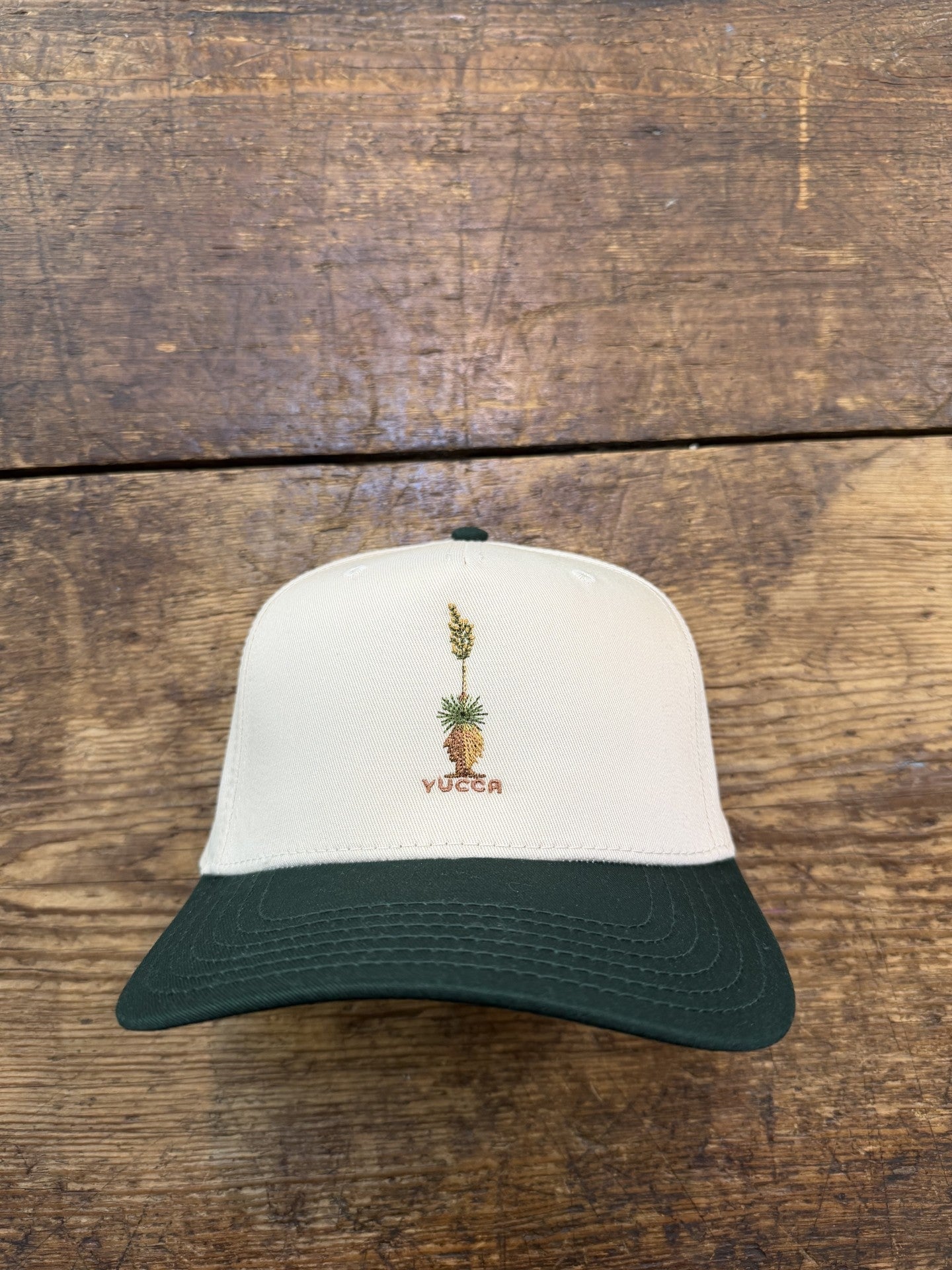 The Shop Hat | Yucca by Ballad of the Bird Dog is a white and dark green baseball cap featuring an embroidered yucca plant and "YUCCA" on the front, shown resting on a wooden surface.