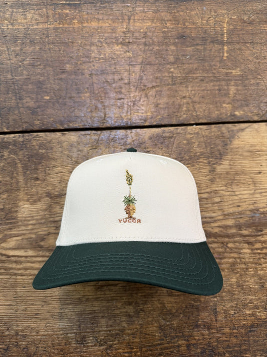 The Shop Hat | Yucca by Ballad of the Bird Dog is a white and dark green baseball cap featuring an embroidered yucca plant and "YUCCA" on the front, shown resting on a wooden surface.