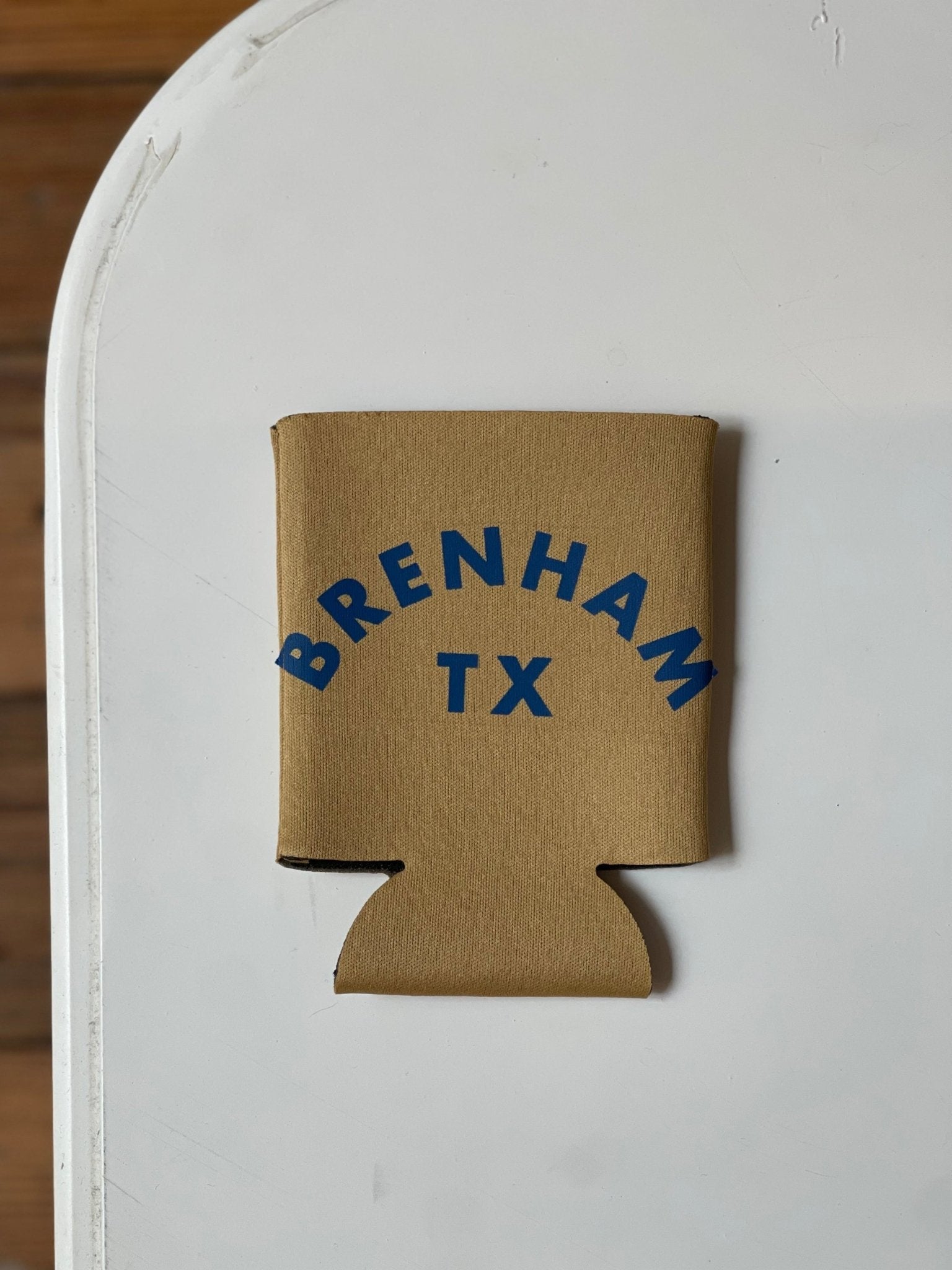 A Ballad of the Bird Dog Shop Koozie featuring "BRENHAM TX" in blue letters rests on a white surface.