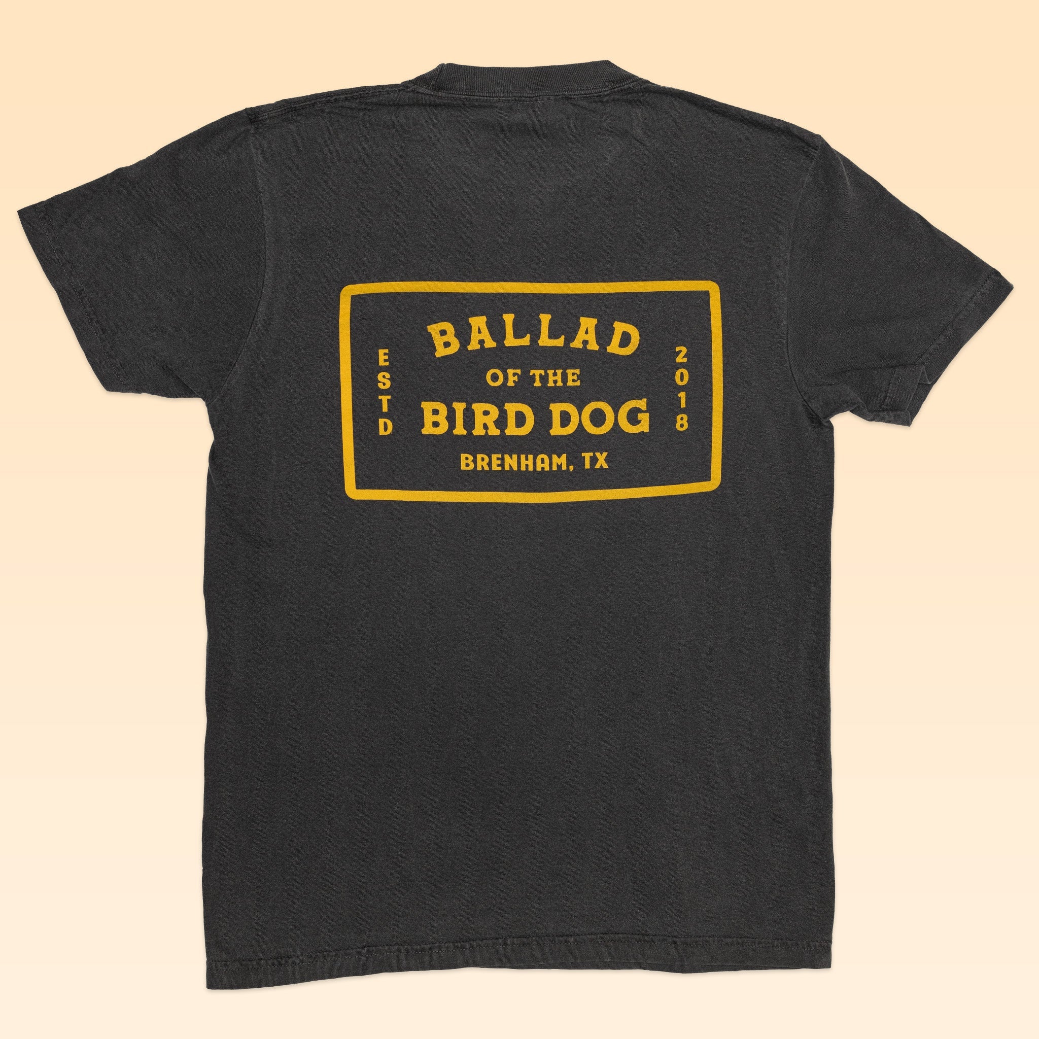 The Ballad of the Bird Dog Shop Shirt features a black vintage design with a yellow flag graphic on the back reading "Ballad of the Bird Dog, ESTD 2018, Brenham, TX," blending heritage style with timeless appeal.