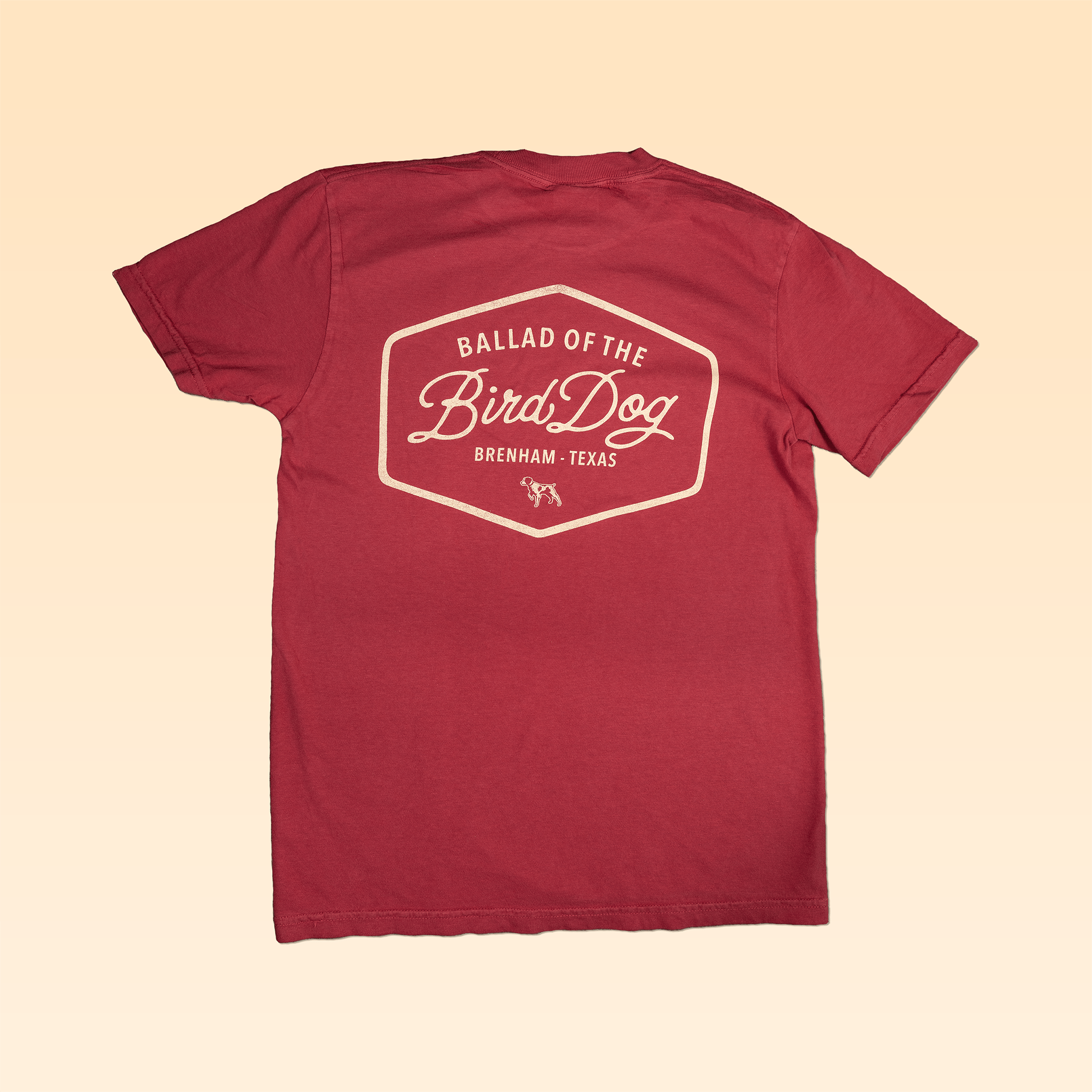 The Shop Shirt | Bird Dog Badge by Ballad of the Bird Dog features a maroon tee with a cream hexagonal back graphic reading "Ballad of the Bird Dog, Brenham, Texas" and a small dog illustration—ideal for vintage hunting tee fans.