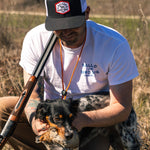 A man in a Ballad of the Bird Dog Shop Shirt with the Bird Dog Classic Shop Logo kneels outdoors with a shotgun, while his hunting dog proudly holds a bird in its mouth.