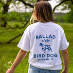 A person wearing a white Shop Shirt | Bird Dog Classic Shop Logo by Ballad of the Bird Dog stands outdoors in a grassy area with trees in the background.