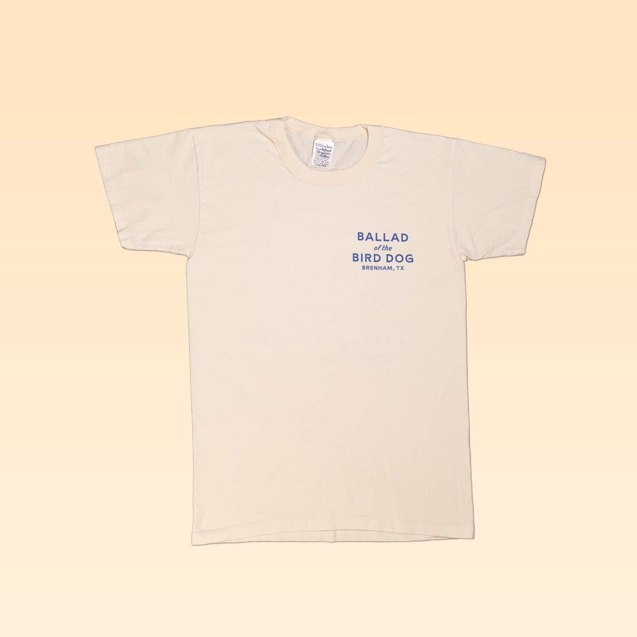 A beige short-sleeve Shop Shirt by Ballad of the Bird Dog, featuring a blue "Ballad of the Bird Dog Brenham, TX" classic shop logo on the upper left chest.