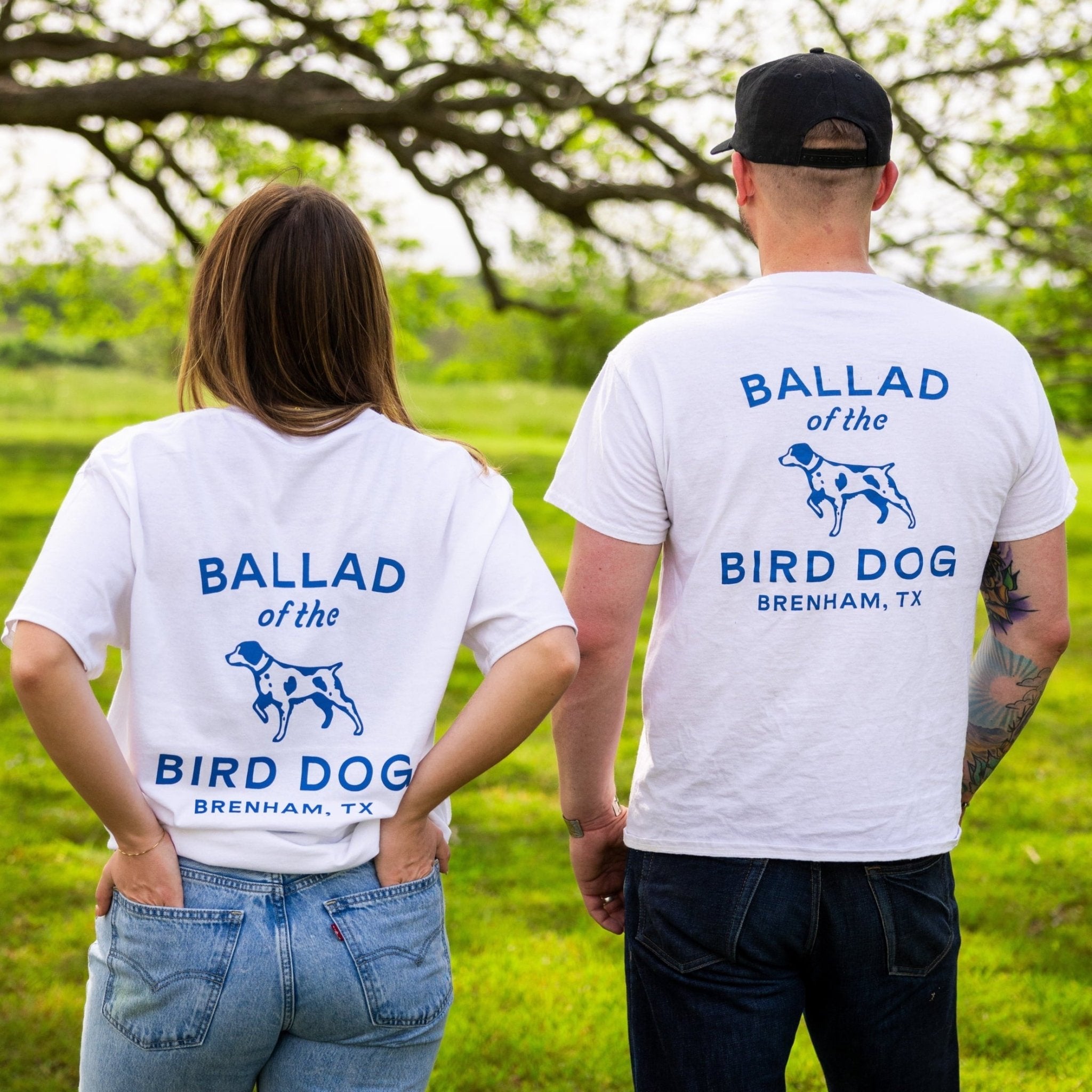 Two people outdoors wear Ballad of the Bird Dog’s Shop Shirt featuring the Classic Shop Logo—an organic cotton tee with a blue dog graphic and "Ballad of the Bird Dog, Brenham, TX" on the back—while facing away from the camera.