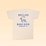 The Shop Shirt | Bird Dog Classic Shop Logo by Ballad of the Bird Dog is a cream-colored tee with blue "Ballad of the Bird Dog Brenham, TX" text and a dog graphic. Made from soft organic cotton for lasting comfort.