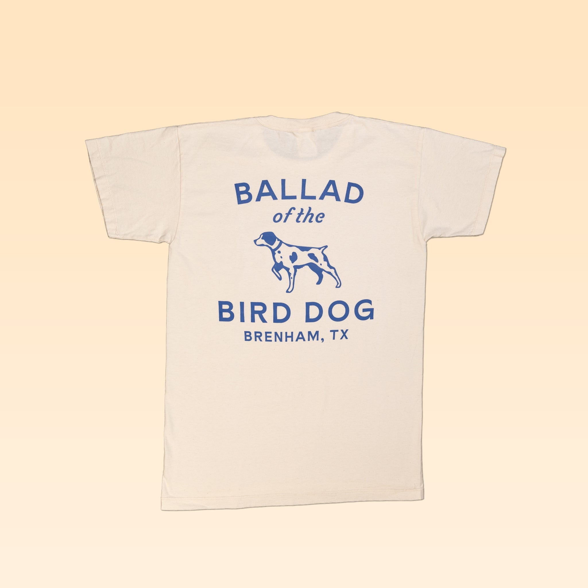 The Ballad of the Bird Dog Shop Shirt features their classic shop logo with a blue dog graphic and "Ballad of the Bird Dog, Brenham, TX" on the back; this cream-colored organic cotton tee is stylish and comfortable.