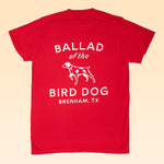 The Ballad of the Bird Dog Shop Shirt features a red design with white text and a dog graphic on the back, inspired by the Classic Shop Logo, reading "BALLAD of the BIRD DOG BRENHAM, TX.