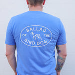 A person wearing the Ballad of the Bird Dog's Shop Shirt | Bird Dog Emblem, featuring "Ballad of the Bird Dog" and a dog graphic on the back, stands against a plain light-colored wall, proudly showing bird dog pride.