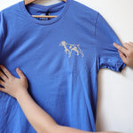 A person holds up the Ballad of the Bird Dog “Shop Shirt | Bird Dog Emblem” on a wooden hanger, featuring a small dog outline on the left chest—a stylish way to show bird dog pride in Downtown Brenham.