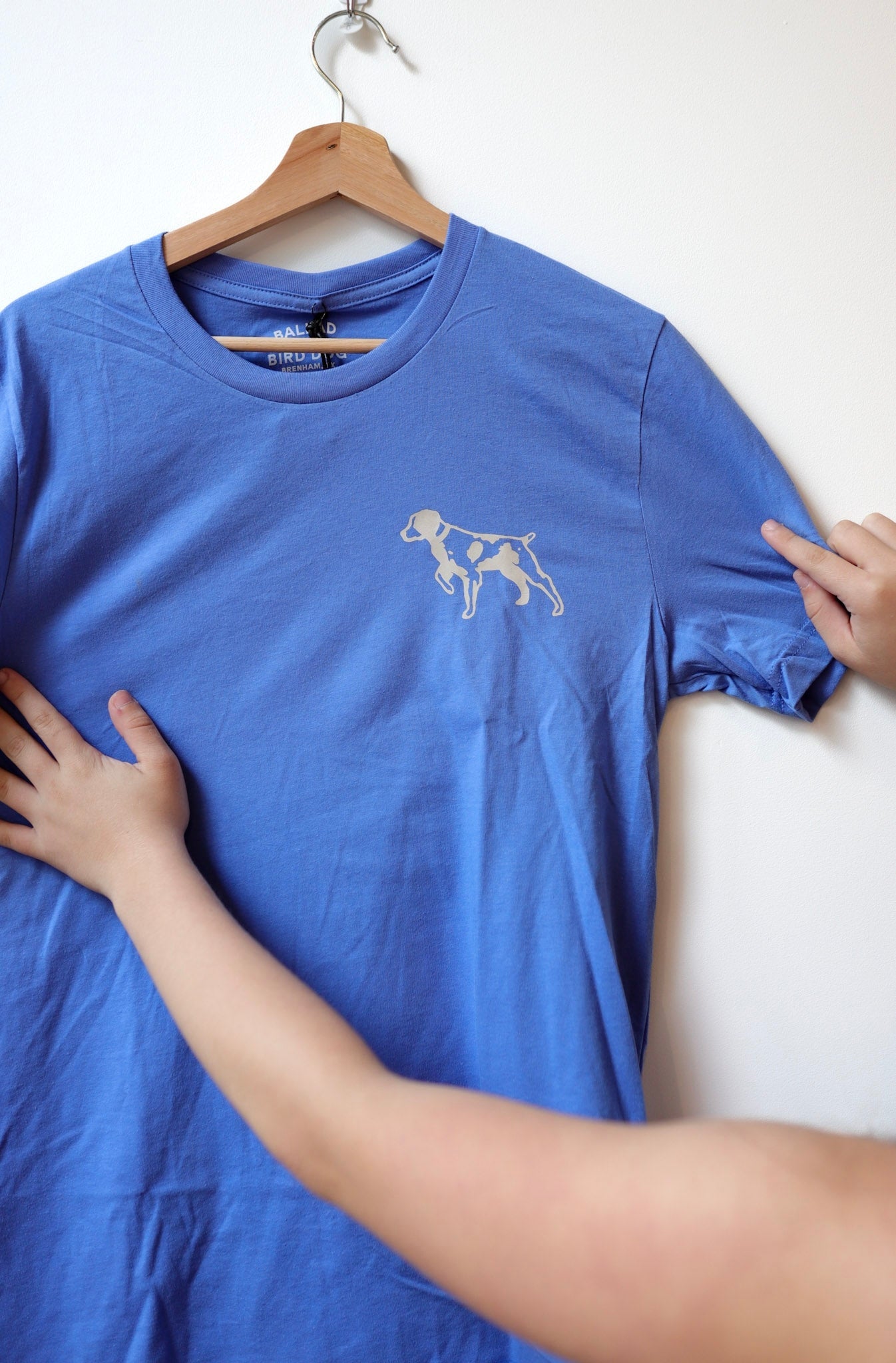 Someone holds the blue Shop Shirt | Bird Dog Emblem by Ballad of the Bird Dog, featuring a small dog outline on the upper left chest, proudly displaying their bird dog pride in Downtown Brenham.
