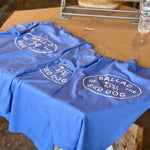 Three blue "Shop Shirt | Bird Dog Emblem | Ballad of the Bird Dog" shirts by Ballad of the Bird Dog are displayed on a table beside a water bottle and hat—perfect for showing your bird dog pride in Downtown Brenham.