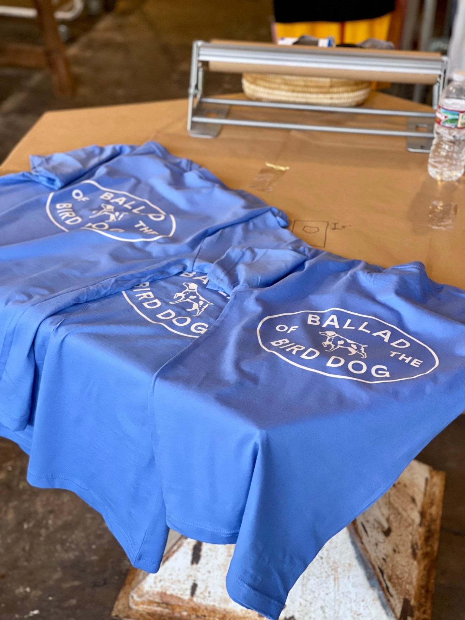 Three blue "Shop Shirts" by Ballad of the Bird Dog, featuring the Bird Dog Emblem and "BALLAD OF THE BIRD DOG," are displayed on a table with a water bottle and hat—perfect for showing your bird dog pride in Downtown Brenham.