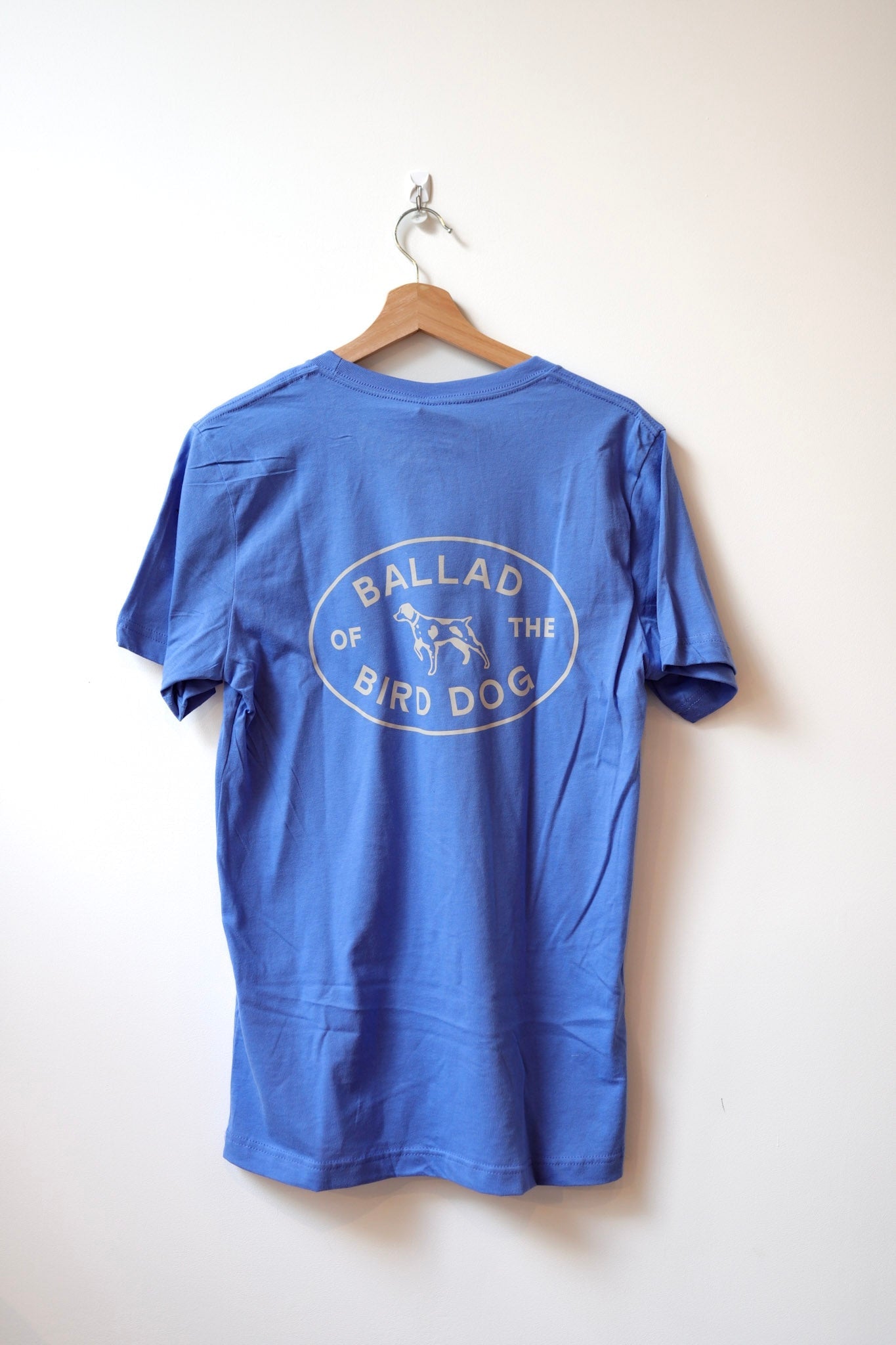 A Shop Shirt | Bird Dog Emblem | Ballad of the Bird Dog by Ballad of the Bird Dog hangs on a wooden hanger, featuring an oval dog logo and "BALLAD OF THE BIRD DOG" text—ideal for showing bird dog pride in Downtown Brenham.