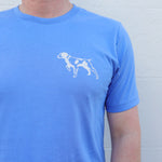 A person wearing the Ballad of the Bird Dog Shop Shirt | Bird Dog Emblem sports a small white dog graphic on the left chest, standing against a light wall and showing off their bird dog pride in Downtown Brenham style.