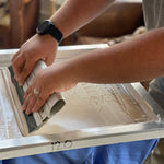 Close-up of a person screen printing a Shop Shirt | Bird Dog Emblem by Ballad of the Bird Dog, spreading ink over a screen on blue cloth, showcasing bird dog pride in Downtown Brenham.