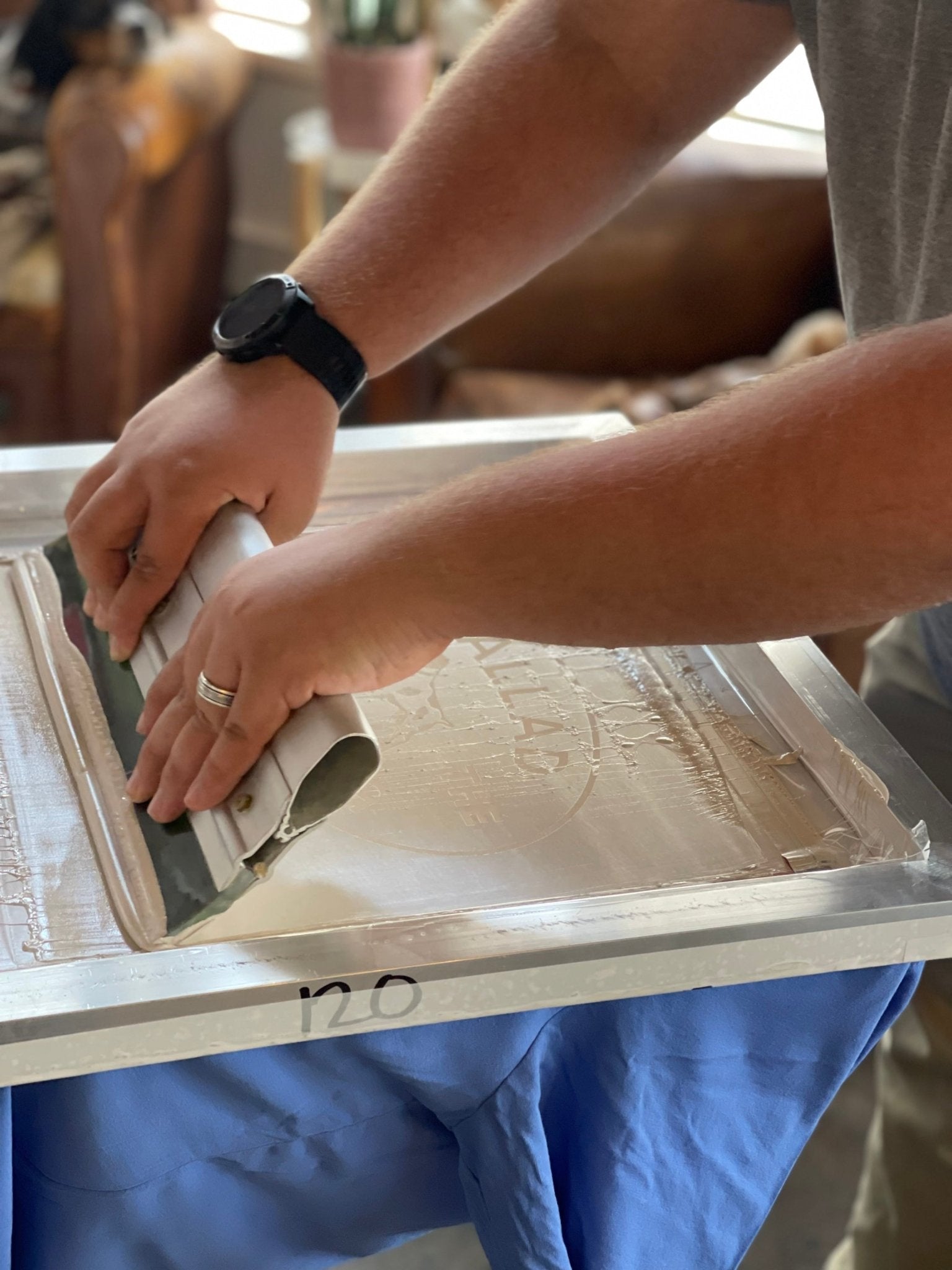 A person uses a squeegee to spread ink across a screen printing frame, preparing to transfer the Ballad of the Bird Dog Shop Shirt featuring the Bird Dog Emblem onto fabric in a Downtown Brenham studio.