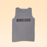 The Ballad of the Bird Dog Honor Tank is a gray sleeveless shirt made from soft garment-dyed fabric, featuring bold black "BIRD DOG" lettering across the chest, shown against a light beige background.