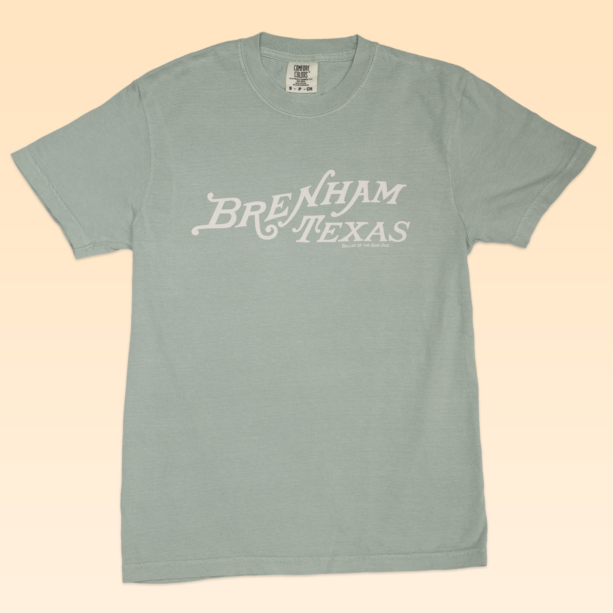 The Shop Shirt | Brenham Texas Heritage Script by Ballad of the Bird Dog features "Brenham Texas" in white script across a light blue shirt, shown on a beige background.