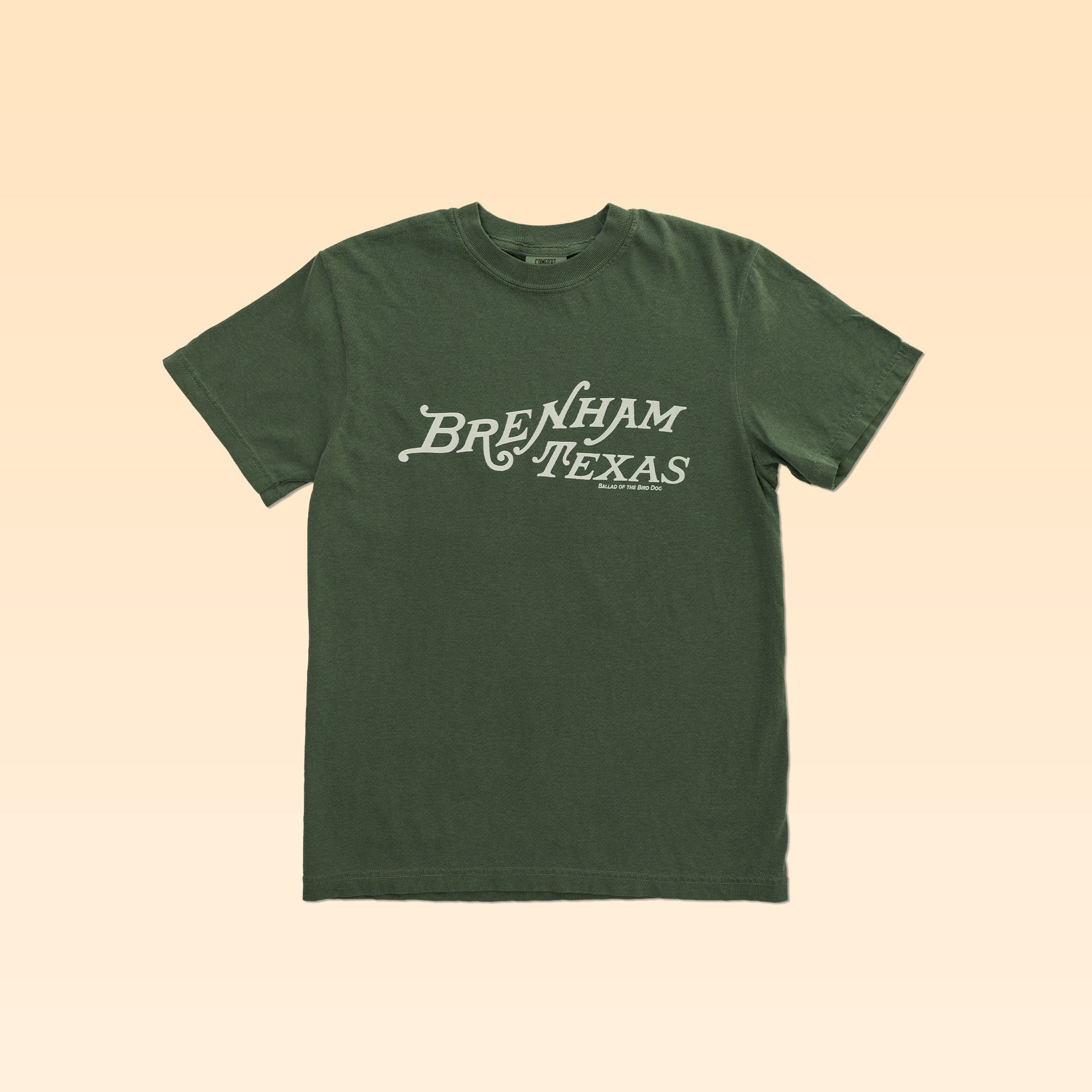 The Shop Shirt by Ballad of the Bird Dog features "Brenham Texas" in white heritage script on green, shown against a beige background—a classic staple for any wardrobe.