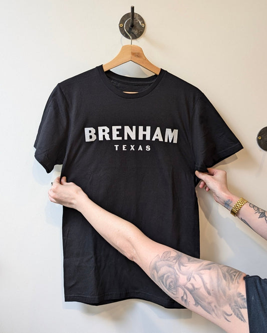 A person holds up the Shop Shirt | Brenham Varsity by Ballad of the Bird Dog, a black shirt on a hanger with "BRENHAM TEXAS" in bold white block letters for a classic collegiate-inspired style.