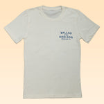 Shop the Ballad of the Bird Dog Circle Clubhouse Tradition Shop Shirt—a white tee with blue "BALLAD of the BIRD DOG BRENHAM, TX" text on the left chest, set against a pale peach background. Perfect for fans of this iconic style!.
