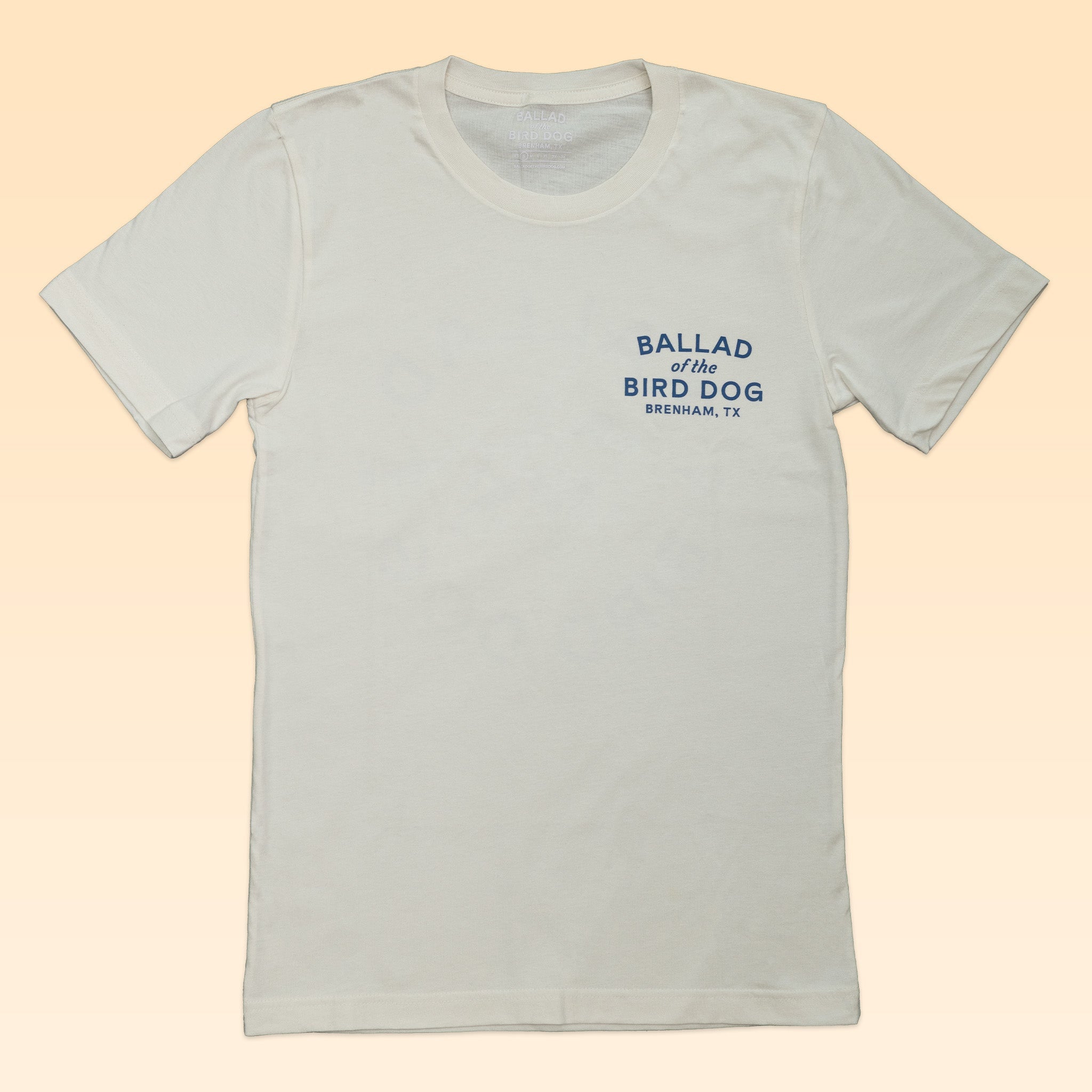 Shop the Ballad of the Bird Dog Circle Clubhouse Tradition Shop Shirt—a white tee with blue "BALLAD of the BIRD DOG BRENHAM, TX" text on the left chest, set against a pale peach background. Perfect for fans of this iconic style!.