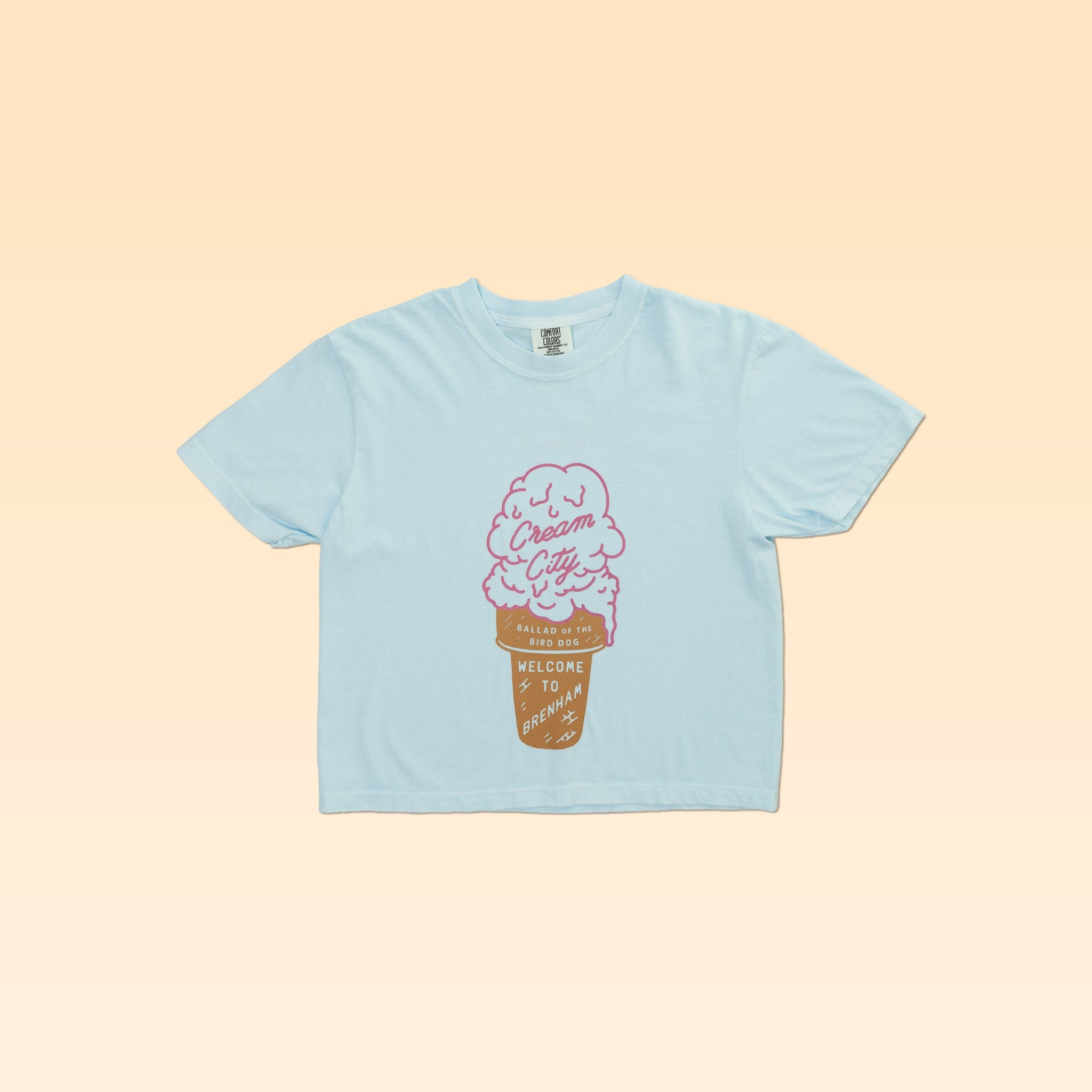 The Ballad of the Bird Dog Shop Shirt | Cream City Crop features a pink ice cream graphic and “Cream City,” “Welcome to Dreamland,” and “Milwaukee, WI” text on a light blue tee, displayed flat on a beige background.