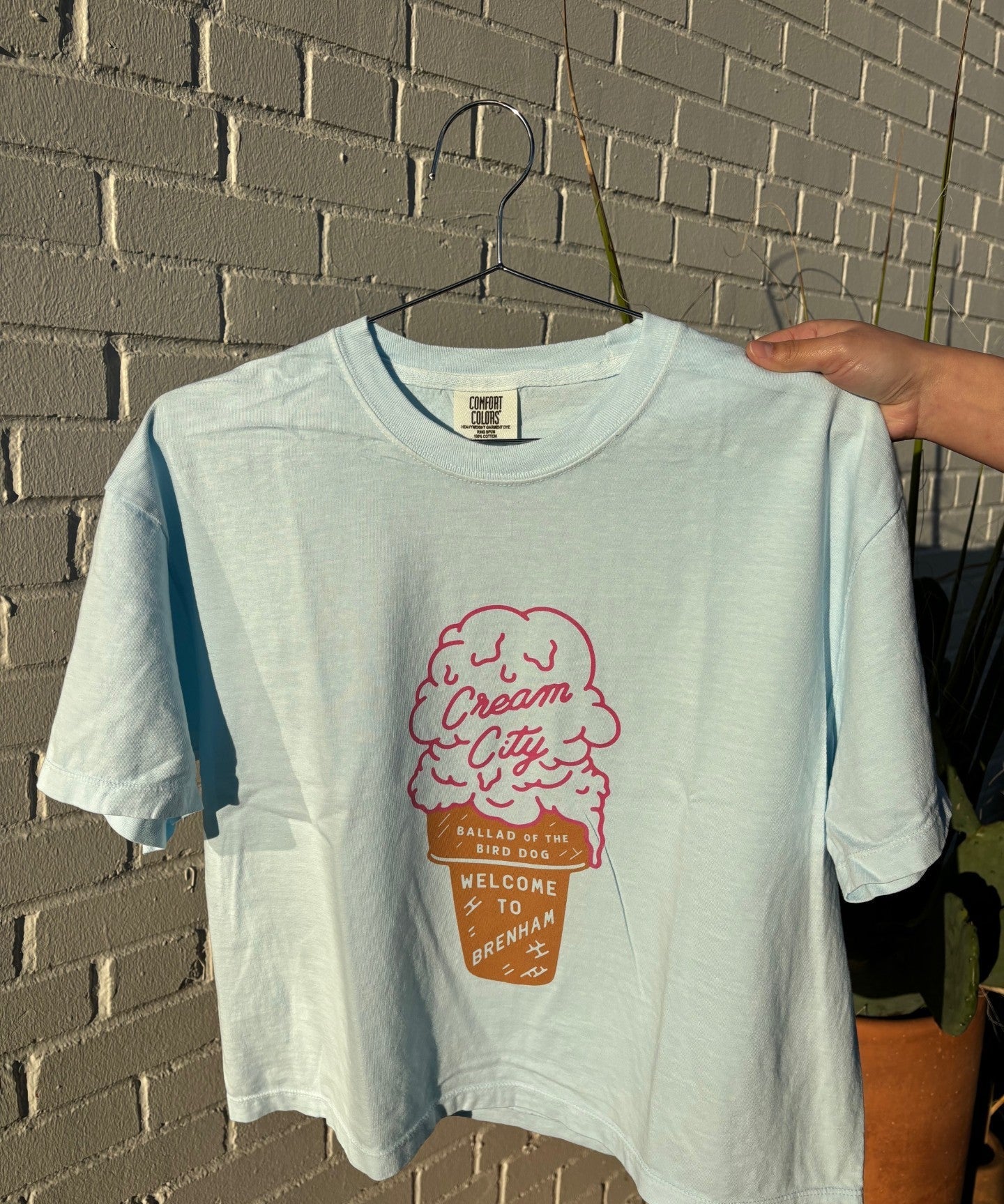 The Ballad of the Bird Dog Shop Shirt | Cream City Crop is a light blue tee with a pink ice cream cone and "Cream City, Welcome to Brenham" on the front, shown held on a hanger against a brick wall.