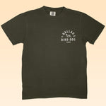 The Shop Shirt | Desert Covey Flush by Ballad of the Bird Dog is an olive green, hand-printed outdoor tee featuring a small white "Ballad of the Bird Dog Texas" graphic with a dog illustration on the left chest.