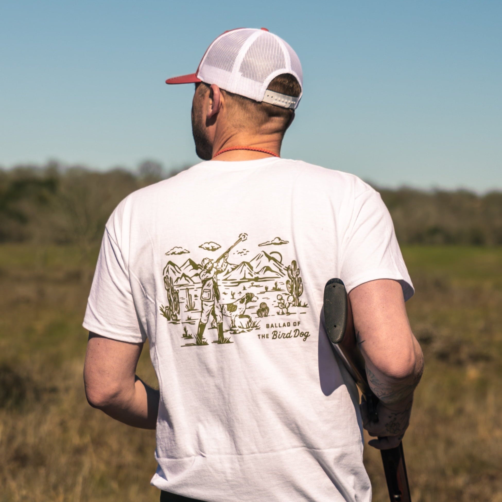 A man in a white Ballad of the Bird Dog Shop Shirt | Desert Hunt and a white mesh cap stands outdoors, holding a shotgun and facing away from the camera.
