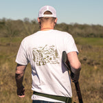 A man in a white Ballad of the Bird Dog Shop Shirt | Desert Hunt and white cap stands outdoors with a shotgun over his shoulder, facing away from the camera in a grassy field.