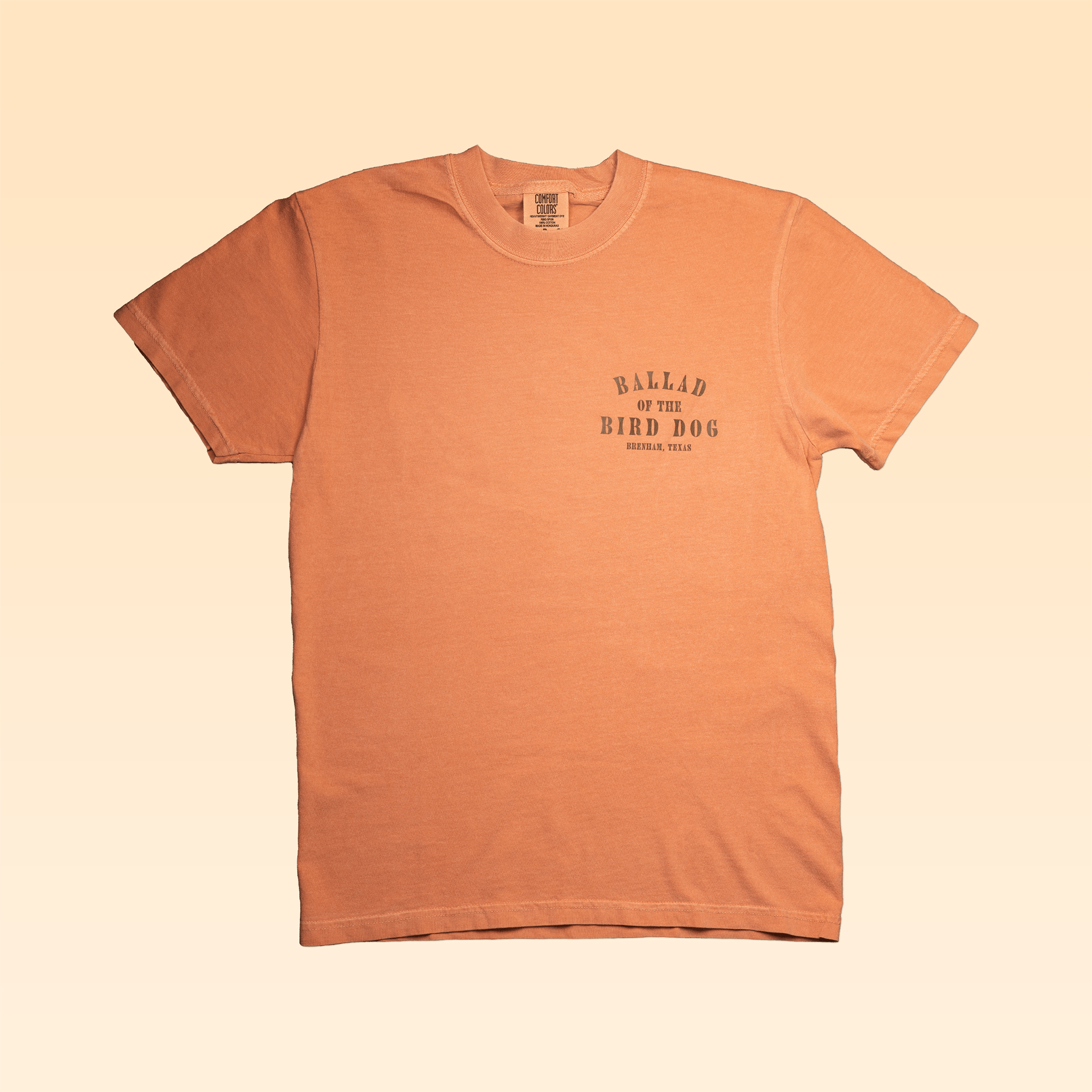 The Shop Shirt | Diamonds by Ballad of the Bird Dog is a peach hand screen-printed shirt featuring “BALLAD OF THE BIRD DOG, BRENHAM, TEXAS” in black on the left chest, displayed on a light gradient background.