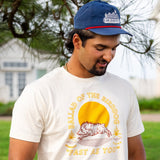A man in a blue cap and Ballad of the Bird Dog Shop Shirt | Fast As You, with a sun and dog design, stands by a tree outdoors, channeling classic country vibes as he looks down.