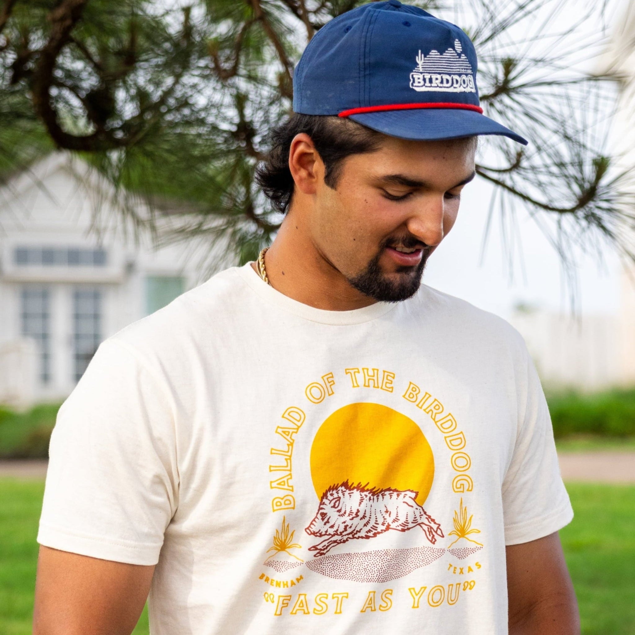 A man in a blue cap and Ballad of the Bird Dog Shop Shirt | Fast As You, with a sun and dog design, stands by a tree outdoors, channeling classic country vibes as he looks down.