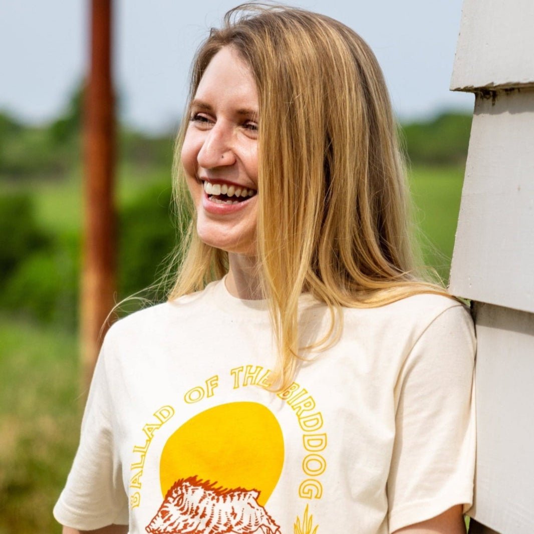 A woman with long blonde hair stands outside, smiling in a Ballad of the Bird Dog "Shop Shirt | Fast As You," posing by a white wooden wall with greenery in the background.