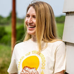 A woman with long blonde hair stands outside, smiling in a Ballad of the Bird Dog "Shop Shirt | Fast As You," posing by a white wooden wall with greenery in the background.