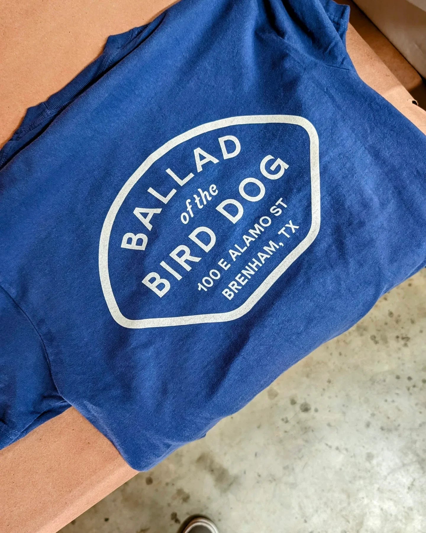 The Shop Shirt | Home Base by Ballad of the Bird Dog is a blue Comfort Colors tee featuring white text: "Ballad of the Bird Dog, 100 E Alamo St, Brenham, TX," displayed on a brown surface.