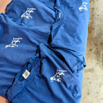 Five blue Shop Shirts | Home Base by Ballad of the Bird Dog, each featuring a white minimalist dog graphic on the left chest, are laid out overlapping on a surface.