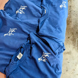Five blue Shop Shirts | Home Base by Ballad of the Bird Dog, each featuring a white minimalist dog graphic on the left chest, are laid out overlapping on a surface.