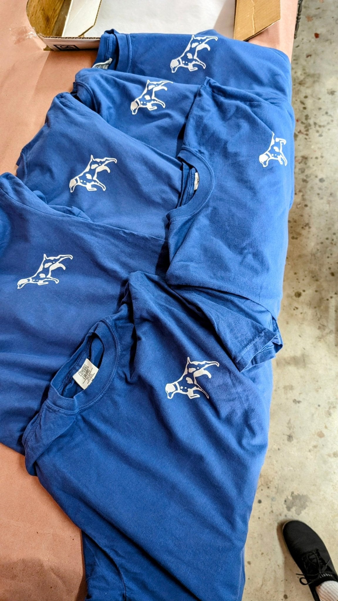 Five blue Shop Shirts | Home Base by Ballad of the Bird Dog, each featuring a white minimalist dog graphic on the left chest, are laid out overlapping on a surface.